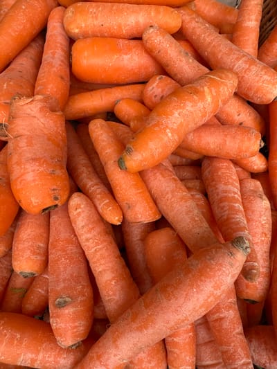 A dense pile of raw, unwashed carrots fills the entire frame. The carrots are predominantly orange, varying in shades, and display irregular shapes and sizes. Visible soil remnants adhere to their surfaces, and some exhibit minor root fragments or superficial blemishes. They are arranged in a disordered, bulk quantity, suggesting storage or display for sale. There are no individuals, other objects, or discernible actions within the scene. The specific location within the city of Wealden, United Kingdom, cannot be determined from the visual information provided.
