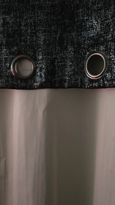 The image displays a close-up view of a dark-colored curtain with two prominent metal grommets on the left side, juxtaposed against a lighter, seemingly plain surface on the right.

The curtain, occupying approximately the left third of the frame, is dark, likely black or a very dark grey, and features a textured, abstract pattern. This pattern consists of numerous small, irregular flecks and streaks of a lighter color, resembling silver or light grey, giving the fabric a distressed or metallic, shimmering appearance. The texture is intricate, creating a visual depth that suggests a woven or printed design.

Two circular metal grommets (eyelets) are visibly embedded into the dark curtain. They are metallic, appearing silver or chrome-colored, with a noticeable sheen reflecting some light. The top grommet is positioned near the upper edge of the curtain, while the bottom grommet is near the lower edge, both aligned vertically. The inner circumference of the grommets appears dark, suggesting the presence of a curtain rod passing through them, or simply shadow within the opening. Subtle reflections are visible on the polished surfaces of the grommets.

To the right of the dark curtain, spanning the majority of the image, is a lighter, uniformly colored surface. This surface appears to be a light grey or off-white, with a faint pinkish undertone that is more apparent in certain areas and crops. It is soft and undulating, with gentle folds and creases visible, suggesting it could be another fabric, a wall with subtle texture, or perhaps a sheet. The lighting on this surface is soft and diffused, creating subtle shifts in tone rather than sharp shadows.

The overall scene is a still life, focusing on the textures and contrasting colors of the materials. No activity or specific event is taking place. The lighting suggests an indoor setting, possibly with ambient light contributing to the soft appearance and the slight pinkish hue on the lighter surface. There is no visible text in the image.