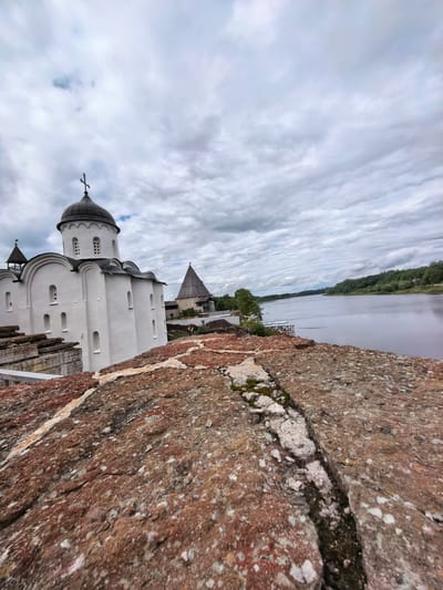 A low-angle, wide-shot photograph captures a view from atop a weathered, reddish-brown stone wall, looking towards an ancient white church with a dark dome and a cross on its peak. The church is situated on a slight elevation, with several arched windows and smaller rectangular openings visible on its facade. To the right of the church, a conical wooden tower stands, part of a larger fortress complex. The scene is dominated by a vast, overcast sky filled with heavy, gray clouds that extend to the horizon. Beyond the fortress, a wide river flows, its surface reflecting the muted sky. The far bank of the river is lined with a dense forest of green trees. The stone wall in the foreground shows signs of age and erosion, with visible cracks and patches of moss. The overall atmosphere is one of historical depth and a somber, cloudy day in Staraya Ladoga, Russia.