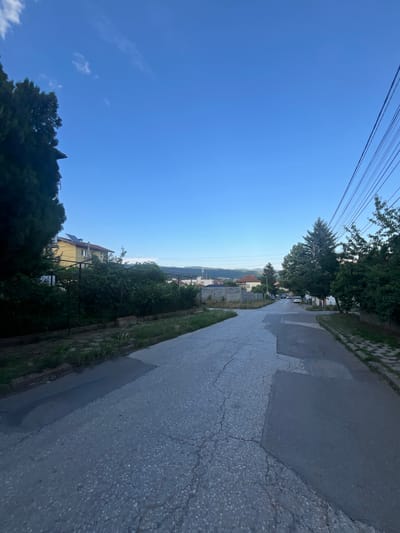 The image depicts a street in Novi Iskar, Bulgaria, during daylight hours. The sky is clear and blue with a few wispy clouds. A paved road, showing signs of wear and repair with patches, dominates the foreground and recedes into the distance. On the left side of the road, there is a tall, dark green coniferous tree and a building with a yellow facade and a red-tiled roof, partially obscured by vegetation. Behind the building and further down the street, more residential buildings are visible, interspersed with trees. Power lines run diagonally across the upper right portion of the image, supported by poles. The street is flanked by greenery, including hedges, trees, and grass. A few parked cars are visible on the road in the middle distance. The overall environment suggests a residential area on the outskirts of a town.