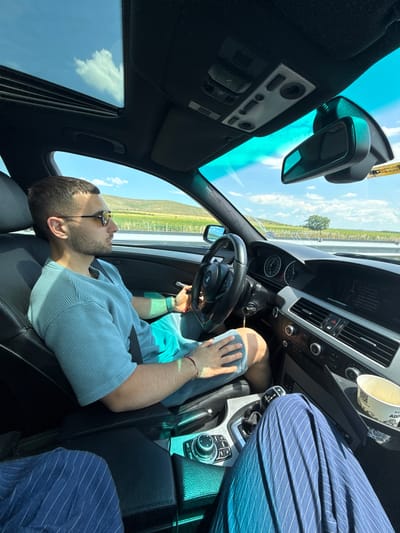 The image depicts the interior of a car on a sunny day. A man with short dark hair, wearing sunglasses, a light blue striped shirt, and shorts is driving. He has a seatbelt fastened. The car's dashboard, steering wheel, and gear shift are visible. Outside the car, a scenic landscape of rolling hills and fields under a bright blue sky with scattered clouds can be seen. The location is identified as Polsko Padarevo, Bulgaria.