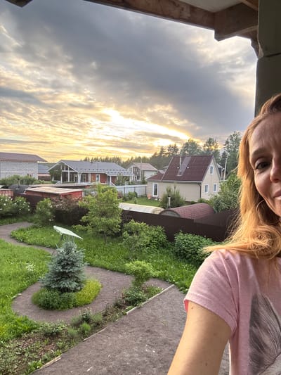 The image depicts a young woman with blonde hair, wearing a pink t-shirt, taking a selfie from an elevated position. She is looking directly at the camera with a slight smile. The background reveals a suburban residential area in the city of Steklyannyi, Russia, characterized by several houses with pitched roofs, some painted red and others beige. The properties are surrounded by green lawns, gardens with various plants, and fences. A small evergreen tree is prominently featured in the foreground, positioned on a curved garden path. The sky above is cloudy with patches of sunlight breaking through, suggesting either dawn or dusk. The overall scene conveys a peaceful, domestic setting.