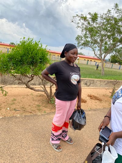 In Vom, Nigeria, a young woman stands outdoors on a paved walkway. She is wearing a black t-shirt and pink checkered pants with red and black accents. She has a black headscarf on and is holding a black plastic bag in her right hand. Her left hand is on her hip. To her right, another person, partially visible, is wearing a white t-shirt and holding two smartphones and a shopping bag. In the background, there is a low building with a red roof, a small tree, and a larger tree. The ground is covered in dirt and sparse grass. The sky is cloudy.