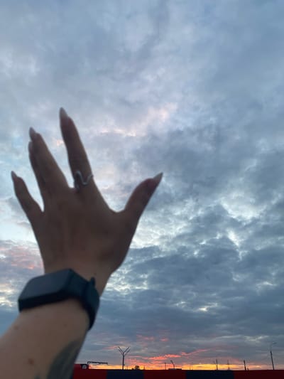 The image captures a close-up of a person's hand, adorned with a ring and a smartwatch, extended towards a cloudy sky. The sky is filled with dramatic clouds, with hints of pink and orange hues visible near the horizon, suggesting either a sunrise or sunset. In the background, a red and black structure runs horizontally across the frame, with streetlights and what appear to be industrial elements visible above it. The presence of a road sign with the number "24" on it is also noticeable. The scene is likely situated in an outdoor urban environment, possibly on the outskirts of Neftekamsk, Russia, given the context of the prompt. The overall mood is one of contemplation or reaching out towards the sky.