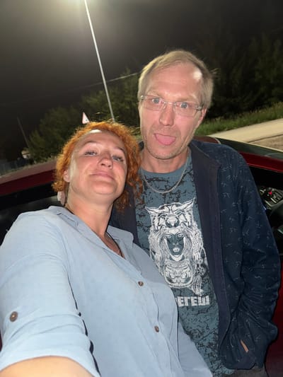 This is a vertical, close-up, selfie-style photo of two adults, a man and a woman, posing closely together. They appear to be in a car at night, with the background showing dark trees and some artificial lighting. The man, on the left, is wearing glasses and a dark blue patterned shirt over a t-shirt with a graphic of a wolf's face. He is sticking his tongue out playfully. The woman, on the right, has red hair and is wearing a light blue button-down shirt. She is smiling and looking towards the camera. The overall mood of the photo is lighthearted and casual, suggesting a fun outing. The image seems to be taken in a rural or semi-rural area given the natural background.