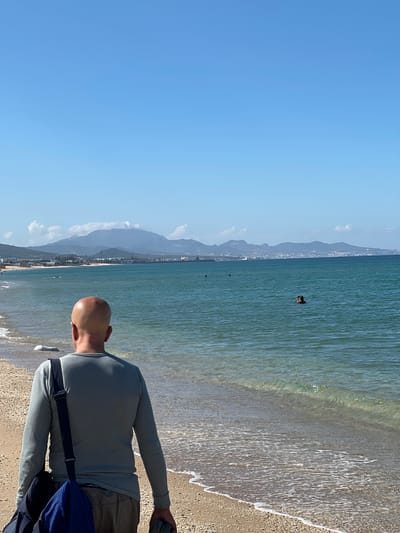 The image shows a beach scene in M'Diq, Morocco. A bald man with a long-sleeved shirt and a blue bag is standing on the sandy beach, looking out towards the sea. The ocean is a turquoise color and small waves are visible near the shoreline. In the distance, there is a coastline with mountains and buildings, indicating the city. There is a person swimming in the water. The sky is clear and blue.
