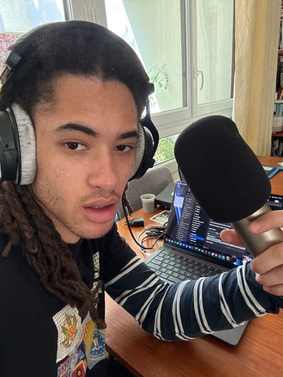 A person is captured in a close-up, rotated selfie, appearing to be in an indoor setting, likely a home office or recording space in Les Ulis, France. The individual, who has dark dreadlocks and stubble, looks directly at the camera with a neutral expression. They are wearing a black long-sleeved shirt with white stripes on the sleeves and a black t-shirt adorned with colorful patches, one of which clearly displays "U.S. DEPARTMENT OF JUSTICE" with an eagle seal. Large, over-ear headphones with light grey earcups are worn on their head.

In their right hand, they hold a silver microphone fitted with a black foam pop filter, pointing it towards themselves, suggesting they are engaged in audio recording or communication. A silver laptop, possibly a MacBook, is open on a wooden desk to their right. Its screen displays a dark-themed interface, possibly code or a terminal window, with various application icons visible on the dock. Another, smaller grey microphone on a stand is visible on the desk behind the primary microphone.

The background features a large window with a white frame and handle, letting in natural light, indicating it is daytime. Outside the window, some greenery is visible. Above the window, light-colored, possibly patterned, blinds are partially drawn. A blurry bookshelf filled with books can be seen in the upper background. Other items on the wooden desk include a white mug and various cables. The overall scene suggests a creative or professional audio setup.