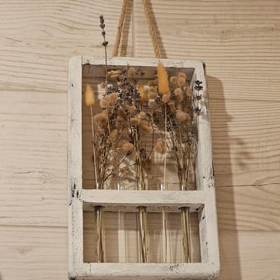 This is a vertical, eye-level, medium shot of a rustic decorative item placed on a light-colored wooden surface. The item is a rectangular wooden frame, painted white with a distressed, chipped finish, which divides into two compartments. The left compartment is filled with dried flowers and grasses, including sprigs of lavender and fluffy seed heads. The right compartment contains several glass test tubes, each holding dried stems or grasses. A thick, twisted rope is attached to the left side of the frame and extends out of the frame to the left, creating a hanging element. The wooden surface beneath appears to be planks with a visible wood grain. The lighting suggests an indoor setting, possibly with natural light casting a shadow from the right side of the frame onto the wooden surface. The overall mood is rustic, natural, and perhaps slightly vintage. No people are visible. There is no visible text. The setting is identified as Vybolovo, Russia.