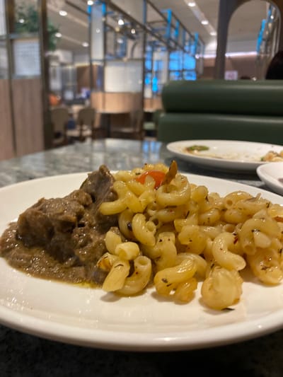 A close-up shot captures a white oval plate holding two distinct food items in a modern dining establishment in Sepang, Malaysia. On the left side of the plate, a generous portion of dark brown, thick, and glossy stew or braised meat is visible, appearing rich and well-cooked. Adjacent to it, on the right, a pile of elbow macaroni pasta, light yellow in color, is seasoned with dark herbs and contains scattered small red flecks, likely chili flakes, and a prominent small red tomato segment. The pasta has a slightly oily sheen.

The background is intentionally blurred, indicating a shallow depth of field, but clearly depicts a spacious, brightly lit restaurant interior. Dark green upholstered booth seating is visible on the right mid-ground. Further back, numerous recessed overhead lights illuminate the area, and distinct blue ambient lighting elements are visible across the far background, possibly part of decorative architectural features or displays. Blurred figures of other patrons and additional tables with white plates, including one partially eaten, can be discerned in the distance. The overall environment suggests a contemporary and upscale dining experience, consistent with a large food court or restaurant within Kuala Lumpur International Airport (KLIA) or KLIA2.