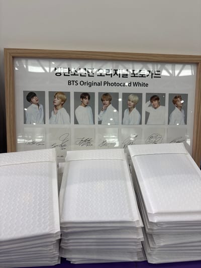 The image shows a framed display of BTS photocards and autographs. The display is on a wall above stacks of white padded envelopes. The text on the display reads "방탄소년단 오리지널 포토카드" which translates to "BTS Original Photocard," followed by "BTS Original Photocard White." There are eight individual photocards of BTS members, each paired with a signature below. The photocards are arranged in two rows of four. The bottom half of the image is filled with stacks of white padded envelopes. The lighting suggests an indoor setting, possibly a retail store or a fan event space. The location in Busan, South Korea, is not provided in the image itself.