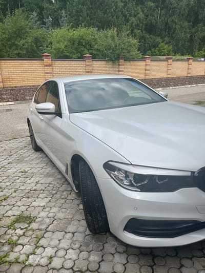 A white BMW sedan is parked on a cobblestone driveway. The car is facing forward, with its front right quarter visible. The driveway is bordered by a low, yellow brick wall topped with decorative brick pillars. Behind the wall, a dense line of green trees forms the background. The sky is overcast, with visible clouds reflecting on the car's hood. The scene appears to be in a residential area.