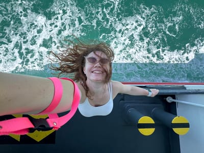 A high-angle, wide shot captures a woman on a boat, with her body positioned vertically in the frame and her head at the top. She is taking a selfie, holding the camera with her left arm outstretched. The woman has reddish-brown hair that is being blown by the wind, and she is wearing sunglasses and a white tank top. She is smiling broadly. Her right arm is extended to her side, and she is leaning against the railing of the boat.
The boat's deck is visible below her, with a black surface that has yellow and black hazard markings. There are two large, cylindrical objects on the deck, painted dark gray with yellow and black circular markings on their ends. To the right of these, a metal railing extends horizontally.
The water surrounding the boat is a vibrant turquoise, with white foam and waves indicating movement. The background consists of the churning sea, with the horizon out of frame. The image appears to have been taken during the daytime, with bright natural light.
The location is described as Unknown, Thailand.