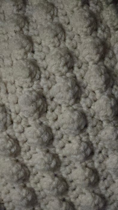 The image depicts a textured surface, possibly a wall or ceiling covered in a bumpy material. The surface appears grainy and uneven, with repetitive, dome-like structures clustered together. The color palette is muted, primarily consisting of shades of gray and beige, giving the impression of a neutral or aged surface. Shadow and light play across the texture, accentuating the three-dimensional aspect of the bumps and creating a pattern of highlights and lowlights.
