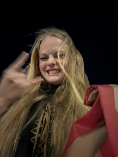 In this medium shot, a young Caucasian female with long, blonde hair is captured against a dark, indeterminate background, possibly indicating an outdoor nighttime setting in Pervomaysky, Russia. She is facing the camera directly, her eyes are closed, and her mouth is open in a wide grin, revealing her teeth. Her left hand is blurred and raised, positioned in front of her face, with the index and middle fingers extended, while her thumb is tucked in. She is wearing a dark garment with a laced front, featuring brown cords woven through eyelets. To the right of the frame, a folded red object, possibly fabric or a bag, is partially visible. The lighting appears to be artificial and from the front, casting minimal shadows. The overall image has a slightly grainy texture and a shallow depth of field, with the background completely out of focus.