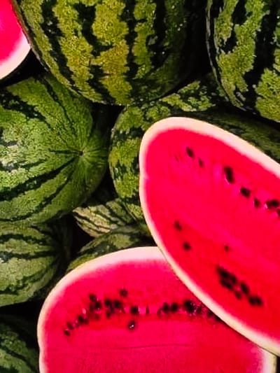 A close-up, overhead shot shows a pile of watermelons. The watermelons are a mix of whole and halved fruits. The whole watermelons have dark green stripes on a lighter green rind. The halved watermelons display bright red flesh speckled with black seeds. The scene appears to be a market stall in Voluntari, Romania, where these watermelons are likely for sale. The lighting is bright, highlighting the vibrant colors of the fruit.