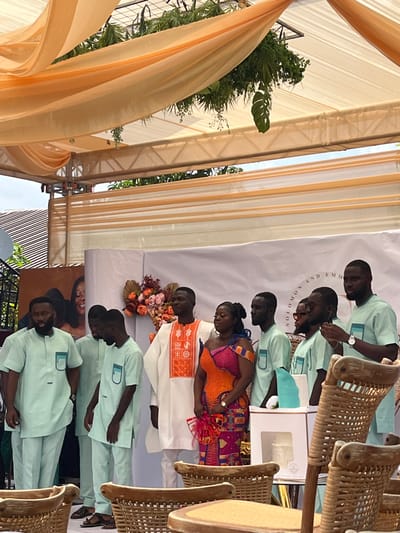 The image depicts an outdoor ceremony in Kumasi, Ghana, taking place under a tent structure. The tent ceiling is draped with light peach fabric and features suspended artificial green foliage, including monstera leaves. Seven individuals of African descent are positioned across the midground.
Central to the group, a man wears a white tunic with orange embroidered patterns and white trousers. To his immediate right, a woman is dressed in a vibrant, multi-colored traditional print gown with an off-the-shoulder design; she holds a small red fan. Five other men wear matching light mint green tunics with dark green pocket details and light mint green trousers. The man on the far left has a beard. The man on the far right is observed checking a watch on his left wrist.
Behind the individuals, a white backdrop is visible, bearing the text "SOLOMON AND EMMA" and an orange and brown floral arrangement. A partial brown panel featuring an image of two smiling individuals is visible on the far left of the background. In the upper left background, a satellite dish and a segment of a tiled roof are present. The foreground contains multiple empty rattan chairs with light-colored seat pads. A small, light blue tiered object rests on a white stand to the right of the central woman.