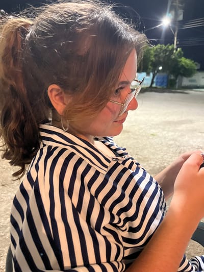 A young woman with brown hair pulled back in a ponytail and wearing glasses and a blue and white striped shirt is seen from the side in Conuco Viejo, Venezuela. She is looking down at her hands, which are holding a cell phone. The background is blurred, but appears to be an outdoor setting with trees and a street light. The lighting suggests it is nighttime.