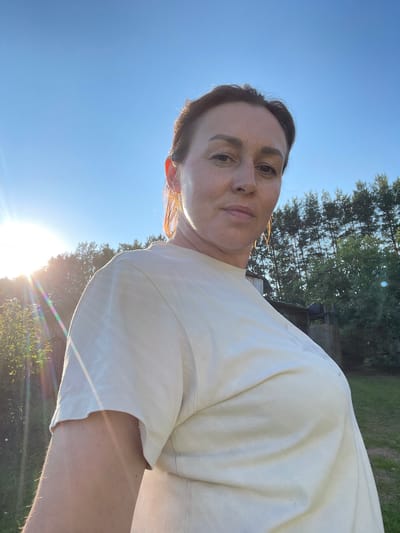 A woman is taking a selfie outdoors in front of a clear blue sky and a line of trees. She is wearing a light-colored t-shirt. The sun is visible on the left side of the frame, casting a flare across the image. The background shows a rural landscape with some wooden structures and green grass. The location is not Bormist, Russia.