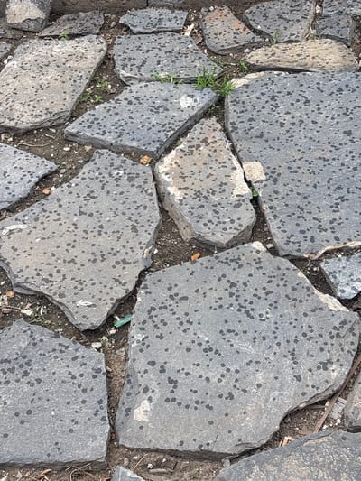 This image depicts an overhead view of an irregularly paved ground surface in Kuklen, Bulgaria. The surface is composed of numerous flat, grey-toned stones of varying polygonal shapes, laid directly on exposed earth.
The stones are generally light to medium grey, with surfaces mottled by numerous small, dark, irregularly shaped spots, consistent with lichen growth or dark mineral inclusions. Some stones exhibit lighter, chalk-like white patches or wear marks. The edges of the stones are irregular and appear to be unpolished or roughly cut.
Gaps between the stones reveal dark brown soil. Within these gaps, small clusters of green grass and other low-lying weeds are visible. Various pieces of debris are scattered across the soil and on the edges of some stones. Identifiable debris includes several off-white cigarette butts, small fragments of green and white plastic, twigs, small stones, and a rusted metallic object near the lower right.
The scene suggests an outdoor, unpaved or poorly maintained pathway or ground area. No individuals, animals, or specific actions are depicted.