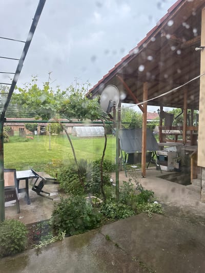The image shows an outdoor scene in Lehchevo, Bulgaria, viewed through a rain-streaked window. Outside, there's a grassy backyard with a white greenhouse in the background. A grapevine grows along a metal structure near the window. A satellite dish is mounted on a pole. A covered outdoor seating area is visible with a stove and table visible through a gap in the covering. A partially covered table and chair set is visible on a paved patio area. There are some plants planted near the patio. The sky is overcast.