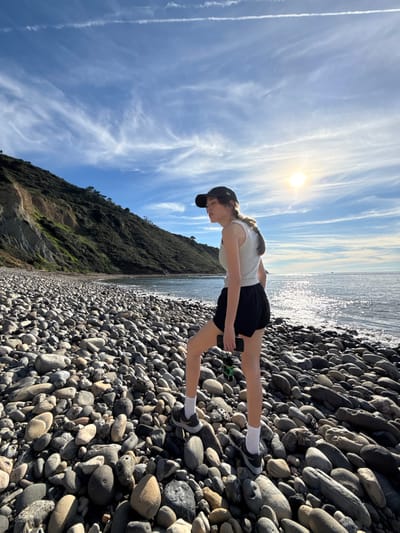 The image depicts a single individual standing on the cobble beach of Lunada Bay in Palos Verdes Estates, United States. The person, appearing female, is positioned slightly right of center, facing towards the right with her head tilted downwards and eyes partially closed, possibly squinting due to the bright sunlight. She wears a dark baseball cap, a light grey sleeveless top, black athletic shorts, white socks, and dark athletic shoes. Her left hand holds a black smartphone with a green attachment. The beach consists entirely of smooth, rounded grey and brown cobbles and pebbles of various sizes. To the left, a steep, vegetated cliff face rises, exhibiting patches of exposed tan earth and dense green foliage. The ocean, calm with bright sun glinting off its surface, extends to the horizon on the right, where a small, indistinct vessel is visible. The sky above is clear blue with sparse, wispy white clouds and a faint contrail. The sun is positioned in the upper right quadrant of the sky.