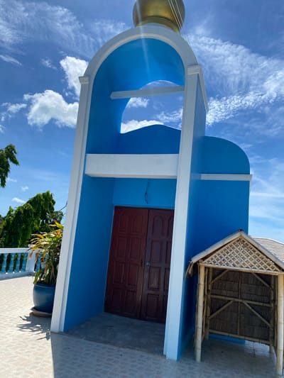 A blue and white structure, likely a bell tower or small chapel, stands against a bright blue sky with scattered white clouds. The structure has an arched opening with a golden dome at the top. To the right of the entrance, there is a small thatched-roof structure made of bamboo. In the foreground, there is a tiled patio. To the left of the main structure, a potted plant sits on the patio, and beyond it, a white railing and lush green trees are visible. The scene appears to be in a tropical environment. The location is identified as Baan Hua Thanon, Thailand. There are no people visible in the image.