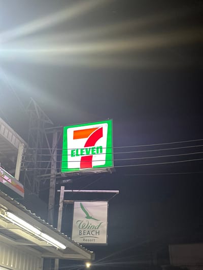 At night, an elevated 7-Eleven sign with a green border and the number "11" in red and orange is prominently displayed. Beside it is a sign for "Wind BEACH Resort" featuring a stylized bird. The signs are mounted on metal scaffolding, and vertical cables run in front of the 7-Eleven sign. The lighting is dim with a lens flare effect.
