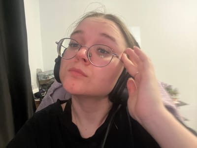 A young female, likely Caucasian, is depicted indoors in Pushchino, Russia. She wears round, thin-framed eyeglasses and dark over-ear headphones. Her hair, appearing dark, is partially visible. She is dressed in a dark, short-sleeved top and a thin silver-colored chain necklace. Her left hand, adorned with a thin ring on the ring finger, is raised to the left side of her face, with fingers near her temple and ear. She exhibits a neutral facial expression while looking directly at the camera. The background consists of a light-colored wall, a dark curtain on the far left, and blurred objects in the mid-ground, including a purple fabric item and other indistinct articles.