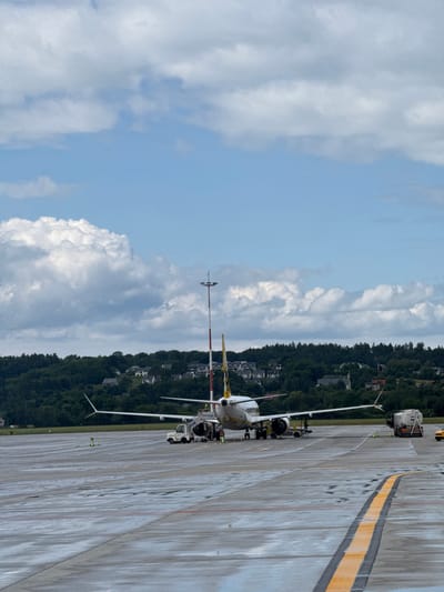 An airplane is parked on the tarmac of the Balice airport, Poland. The aircraft, likely a Boeing 737 based on its size and engine placement, is white with yellow and black accents on its tail. A set of mobile stairs is connected to the front passenger door, and several ground crew members are visible around the plane. A small service vehicle, possibly a baggage tug, is positioned near the stairs. Further down the tarmac, a tanker truck is parked, likely for refueling the aircraft, and a yellow taxi or service vehicle is also visible. The ground appears wet, suggesting recent rain. In the background, a dense, tree-covered hillside rises, dotted with houses, indicating the airport is situated near a residential area. The sky is a bright blue, with scattered white, fluffy clouds. A tall red and white striped pole with lights is also present on the tarmac.