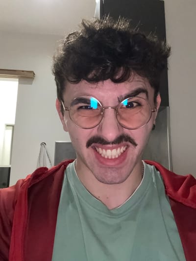 A young man with dark curly hair, light skin, and round glasses smiles broadly at the camera. He appears to have drawn a large mustache on his face. He wears a light green t-shirt and a red jacket. The background is a neutral wall and part of a doorway. The image is rotated sideways.