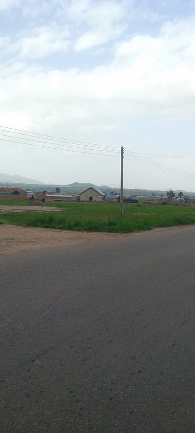 In the city of DU, Nigeria, a road runs through a rural or semi-rural area. The road occupies the foreground of the image, with a grassy verge separating it from a construction site or undeveloped land in the middle ground. In the middle ground, there is a building with a light-colored, sloped roof, and several piles of material, possibly dirt or building supplies, are visible. A single utility pole stands to the right of the building, with power lines stretching across the scene horizontally. In the background, rolling hills form a distant horizon under a cloudy sky. A single individual is visible walking on the grassy area near the construction site, facing away from the camera. The overall scene depicts a developing area with evidence of construction and infrastructure in a natural landscape.