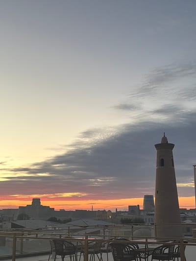 This photograph captures a sunset over the historic city of Khiva, Uzbekistan, from a rooftop vantage point. In the foreground, a glass-railed balcony is furnished with several dark-colored wicker chairs and tables. The middle ground is dominated by the silhouette of the Ichon-Qala, Khiva's walled inner city, with its distinctive fortress-like structures and minarets. A tall, slender minaret, likely the Kalta Minor or another prominent tower, stands prominently on the right side of the frame. The sky is a canvas of warm sunset hues, with streaks of pink, orange, and yellow blending into the soft grey of the clouds. In the distance, the faint outlines of more buildings and perhaps a Ferris wheel suggest the urban landscape beyond the ancient walls. The overall impression is one of serene beauty and historical grandeur as day transitions into night.