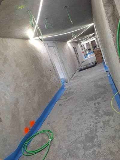 The image depicts an interior construction site, possibly a hallway or corridor, in Jette, Belgium. The scene is dominated by unfinished concrete walls and ceiling. Bright, linear light sources are visible along the ceiling, creating sharp shadows. Green plastic conduits are attached to the ceiling and hang down in several places. The floor is a rough concrete surface with numerous footprints, suggesting recent activity. Blue protective sheeting is laid along the base of the walls. In the middle ground, a cardboard box labeled "Kingspan Kooltherm" is present, along with two rolls of dark material, possibly insulation or protective sheeting. A coil of green tubing is also visible on the floor in the foreground. The overall impression is one of ongoing construction and preparation for finishing work.