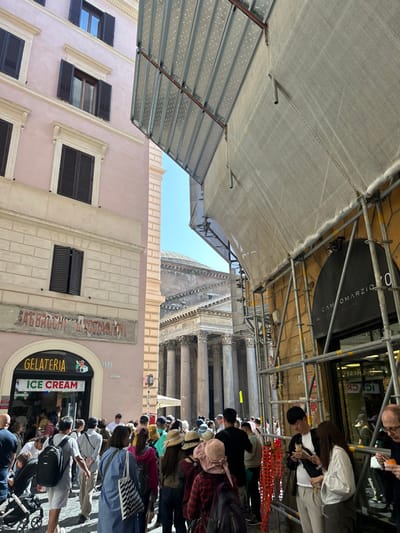 The image shows a crowd of people standing on the street in front of the Pantheon in Rome, Italy. The coordinates provided place the scene in the vicinity of the Pantheon, a famous Roman temple. The Pantheon, partially obscured by scaffolding, is visible in the background. The scene is framed by two buildings, one of which is under construction, with scaffolding covering its facade. The scaffolding is covered with a white tarp, which is partially obscuring the view of the Pantheon. The building on the left side of the image is painted pink and features a storefront for an ice cream shop. A group of people are gathered in front of the ice cream shop, while a larger crowd is gathered in front of the Pantheon. Several people are wearing hats, and some are holding bags or backpacks. Some people are looking at their phones, and some are talking to each other. The street is paved with cobblestones. The weather is sunny and bright.