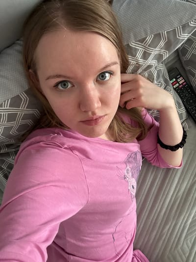 An individual, identified as an adult female, is depicted in a close-up, high-angle shot, lying on her back. She possesses light brown hair, centrally parted, which frames her face and extends onto the bedding. Her eyes are blue with visible black eyeliner on the upper lash line. She exhibits a fair complexion and a neutral facial expression, with her gaze directed towards the camera.
She is wearing a long-sleeved pink top. The top features a graphic design on the left chest area, illustrating a stylized profile of a head with flowing hair and floral elements. A black scrunchie is present on her right wrist. Her left hand is partially visible, resting near her left temple or ear.
The subject is positioned on bedding. Behind her head is a pillow with a grey and white geometric pattern. To her right, a portion of a grey duvet cover or sheet with horizontal stitched lines is visible. A black television remote control, displaying multiple colored buttons (red, green, yellow, blue) and white text including "DVD," is located on the bedding near her head. The environment appears to be an indoor setting.
The specific location in the city of Nizhnekamsk, Russia, cannot be determined from the visual information provided in the image.
