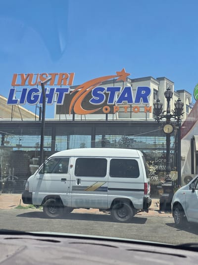The image depicts a street scene in Bukhara, Uzbekistan. A white van, identified by the "DAMAS" badging, is parked on the right side of the frame. To the right of the van, a white car is partially visible. Above and behind the vehicles, a building features a prominent sign with orange and blue lettering that reads "LYUSTRI LIGHT STAR OPTOM". The building appears to be a commercial establishment, with large glass windows displaying merchandise, including decorative lights and what seem to be floral arrangements. A vintage-style street lamp with multiple globes and a clock is situated in front of the building, to the right of the van. The sky is clear blue, indicating a sunny day. The overall impression is that of a commercial district in a city.