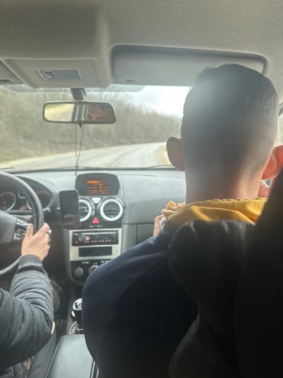 The image shows the interior of a vehicle in motion, traveling on a winding asphalt road in Gorna Rositsa, Bulgaria. The perspective is from a rear passenger seat looking forward. A male driver is partially visible on the left, with both hands gripping a black steering wheel. He wears a dark, quilted jacket, and a male face, presumed to be his, is reflected in the rearview mirror. In the front passenger seat, a male passenger is visible from behind, wearing a dark blue long-sleeved garment over a yellow layer, actively eating a sandwich or similar food item held in his hands.
The dashboard features a speedometer on the left and a tachometer on the right. The center console includes air vents with chrome rings, a red triangular hazard light button, and an aftermarket car radio unit. A black smartphone is mounted vertically on the dashboard to the left of the center console. A small crucifix hangs from the rearview mirror. The vehicle appears to be a left-hand drive with a manual transmission, as indicated by the gear stick visible between the front seats. The exterior environment, viewed through the windshield, shows a two-lane asphalt road curving to the right, bordered by bare trees and sparse vegetation under an overcast sky.