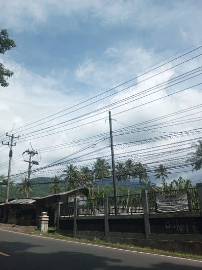 A landscape scene features a road and a low wall beside it. Behind the wall, there is a building under a sloping roof and palm trees. Mountains can be seen in the distance. Overhead are numerous power lines crisscrossing the sky. The sky is partly cloudy with a mix of blue sky and light, white clouds. A utility pole stands near the building.