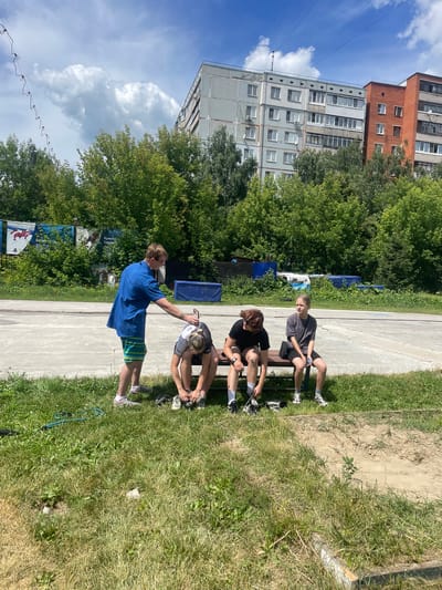 A group of four individuals are gathered outdoors on a sunny day. Three young people are seated on a dark-colored bench, each in athletic attire, engaged in lacing up their shoes. One person, a young female with reddish-brown hair, is focused on her footwear, while the other two, a young female with blonde hair and a young male, are also attending to their shoes.
A fourth individual, a young male wearing a bright blue short-sleeved shirt and striped shorts, stands to the left of the bench. He is interacting with one of the seated individuals, possibly assisting them with their shoes, as he holds a black object.
The scene appears to be an outdoor recreational area, likely a sports ground, indicated by the concrete surface and the surrounding grassy areas. In the background, dense green trees line the perimeter, beyond which are multi-story apartment buildings. The architecture of the buildings suggests a Soviet-era construction, common in many Russian cities. Banners with Cyrillic text are visible on a fence in the mid-ground. The sky is blue with scattered white clouds. The overall impression is a casual, outdoor activity taking place in a residential area of Tula, Russia.