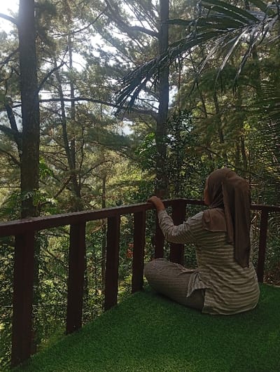 The image depicts an elevated outdoor platform overlooking a dense forest, identified as being located in Kajar, Indonesia. An adult individual, viewed from behind, is seated cross-legged on a green artificial turf surface that covers the platform's floor. The individual wears a light-colored, horizontally striped long-sleeved tunic, dark textured lower garment, and a solid brown hijab covering the head and shoulders. Their left hand rests on the top rail of a dark brown wooden railing that extends across the foreground. Beyond the railing, a dense forest of tall conifer-type trees, with visible trunks and green foliage, extends into the background. Some large, dark green palm-like fronds are visible on the right side of the frame. The scene is illuminated by natural daylight filtering through the tree canopy.