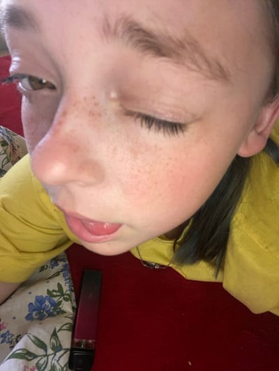 The image shows a close-up, low-angle shot of a person's face, with their head tilted. They have fair skin with freckles across their nose and cheeks. Their eyes are closed, and their eyelashes are dark and prominent. The person's tongue is slightly extended, and their lips are a light pink color. Dark hair is visible, some of which appears to be pulled back. The person is wearing a yellow garment, possibly a hoodie or sweater.
In the background, a red surface is visible, which could be a rug or carpet. To the right, a dark red, cylindrical object with a black cap is lying on a white fabric with a floral pattern featuring blue flowers and green leaves. The lighting suggests an indoor setting, possibly with natural light. No specific time of day or weather conditions can be determined. There is no visible text in the image. The setting is identified as Nova Praha, Ukraine. The activity appears to be a moment of rest or relaxation.