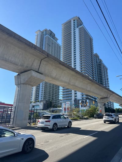 The image shows a street scene in Burnaby, Canada, under a clear blue sky. In the foreground, a concrete elevated structure, likely part of a transit line, dominates the left side of the frame, casting a long shadow across the asphalt road. Several cars are parked and driving on the road. A white Audi SUV is parked on the right side of the road, and a white Tesla is partially visible in the lower right corner, facing away from the viewer.

In the background, several modern high-rise buildings with a distinct architectural style featuring numerous balconies are visible. Some of these buildings have a stepped, tiered design. To the right of the elevated structure, traffic lights and street signs are visible. One sign reads "THE OF LOUGHEED" and another shows "Austin St." A gas station with multiple pumps and branding, including "Esso," is also present. The scene appears to be set during the daytime, given the bright sunlight and clear sky, suggesting fair weather. No people are visible in the image.