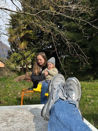 The image depicts an outdoor scene in Erba, Italy, under natural daylight. The foreground presents the lower legs and feet of an individual, clad in blue denim jeans and silver-grey athletic sneakers, resting horizontally on a light-colored, textured surface. In the midground, an adult female with long brown hair, wearing a black jacket and blue jeans, is seated on an orange metal slatted chair. She holds an infant, who is wearing a grey knit beanie and a light olive-green padded jacket, and holds a white object to its mouth. A pair of bright yellow children's rain boots rests on the orange chair beside the infant. The background displays a grassy area with scattered yellow dandelions, dense green foliage including large evergreen trees, and deciduous trees with visible early spring buds. A portion of a building is visible on the far left horizon.