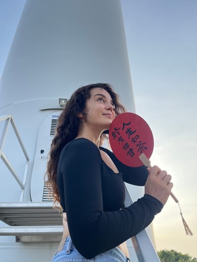 A young woman with long, curly dark hair stands on a metal staircase in front of a large, white, cylindrical structure, possibly a wind turbine tower, in Mui Ne, Vietnam. She is wearing a black, long-sleeved top and light blue denim jeans. She is holding a red, circular fan with black Chinese characters written on it. The fan is positioned in front of her face, and she is looking up and to her right, towards the sky. The sky is a clear, pale blue, indicating it is daytime. In the background, a hint of green foliage can be seen at the bottom right corner of the image. The overall lighting suggests it is late afternoon or early morning.