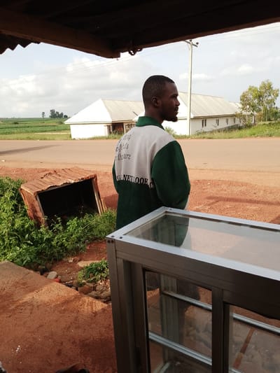 A man is standing outside, his back to the camera, looking to the right. He is wearing a green and white jacket with the words "GENE LOOK" printed on the back in large, dark letters, and smaller text above it. He has short, dark hair and a beard. The man appears to be in a rural or semi-rural setting, with a dirt road in front of him and fields in the background. To his left is a dilapidated, rusted metal structure, possibly a shelter or bin, surrounded by green vegetation. In the background, there are several white buildings with metal roofs, and further in the distance, lush green fields and trees. A power line pole and wires are visible in the sky. The lighting suggests it is daytime, likely late afternoon, with a bright but slightly overcast sky. The overall scene is somewhat rustic and suggests a modest, outdoor commercial or agricultural environment. The man's posture is relaxed, and he appears to be observing something off-camera.