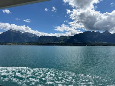 This image depicts a scenic view from a boat on Lake Thun, looking towards the mountains behind Hünibach, Switzerland. The foreground features the turbulent white wake of the boat on the deep blue water. In the middle ground, the calm surface of the lake stretches towards a distant shoreline dotted with trees and buildings. A single white sailboat is visible in the distance on the left side of the lake. The background is dominated by a range of majestic, dark green mountains, some with snow-capped peaks, under a bright blue sky filled with fluffy white clouds. The overall scene is one of natural beauty and tranquility, likely captured on a sunny day.