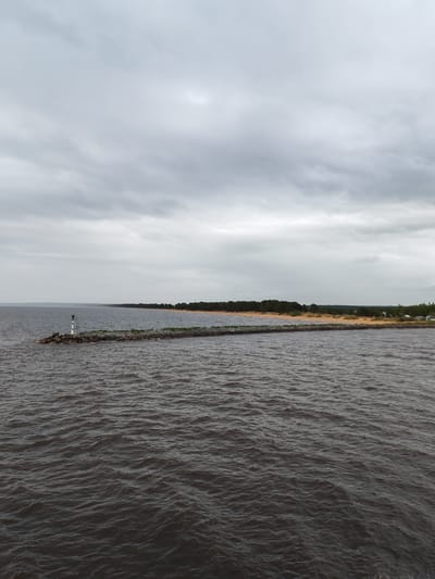 The image depicts a seascape under an overcast sky, possibly in the vicinity of Ozernoe Ust'e, Russia. In the foreground, dark, choppy water stretches towards a long, rocky breakwater that extends into the sea. The breakwater is topped with sparse green vegetation and a white and black navigational marker is at its end. Beyond the breakwater, a shoreline of sandy beach is backed by a dense band of coniferous trees. A few buildings are visible in the distance.