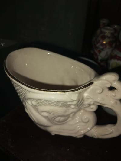 A ceramic cup with a dragon's head design for the handle sits on a dark surface. The cup is off-white with a gold rim. The dragon's head is intricately sculpted with visible scales and facial features. In the background, out of focus, are decorative ceramic vases with floral patterns. The lighting is dim, emphasizing the textures and details of the cup's design.