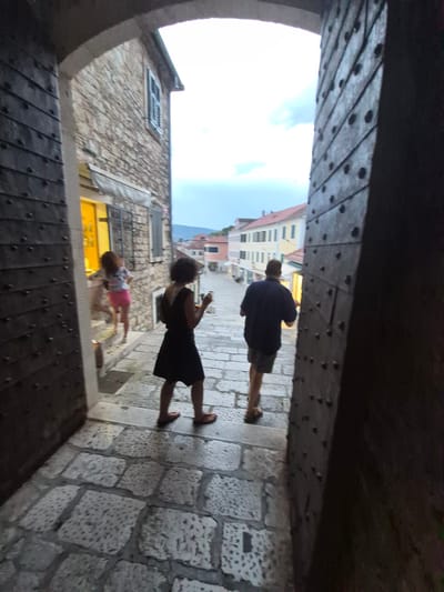 The image depicts three people walking through a stone archway, possibly an entrance to a historic town. The archway is made of heavy, dark wood with visible rivets. The ground is paved with cobblestones.
In the foreground, on the right, a woman in a black dress is walking away from the camera, holding a phone in her hand. To her left and slightly further ahead, a young child in a pink skirt and a patterned top is also walking away from the camera. Further ahead, a man in a dark shirt and shorts is walking away, also looking at his phone.
Beyond the archway, buildings with stone facades and tiled roofs are visible, suggesting an old European town setting. The sky is bright and appears to be daytime, possibly late afternoon given the angle of light and shadows. The overall scene suggests a casual stroll through a picturesque location. The location context provided is Herceg Novi, Montenegro. There is no visible text in the image.