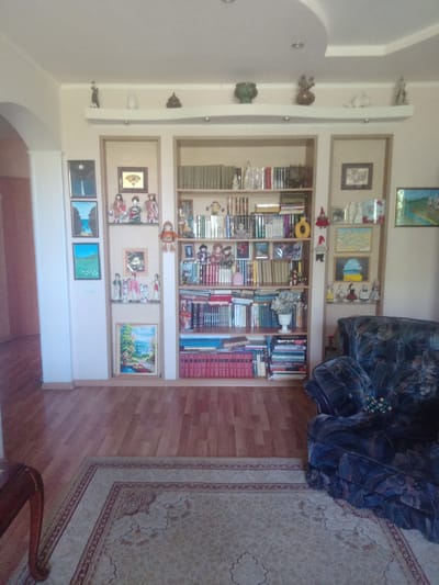 The image shows the interior of a room, likely a living room or study, in an apartment or house located in Oskemen, Kazakhstan. The room features a large built-in bookshelf filled with books, decorative items, and framed pictures. The bookshelf is the central focus, displaying various volumes, figurines, and artwork. To the left of the bookshelf, there is a white wall with several framed pictures arranged vertically. In the foreground, a patterned rug covers a portion of the wooden floor. To the right of the bookshelf, a dark-colored armchair is visible. The overall impression is of a well-decorated and organized living space.